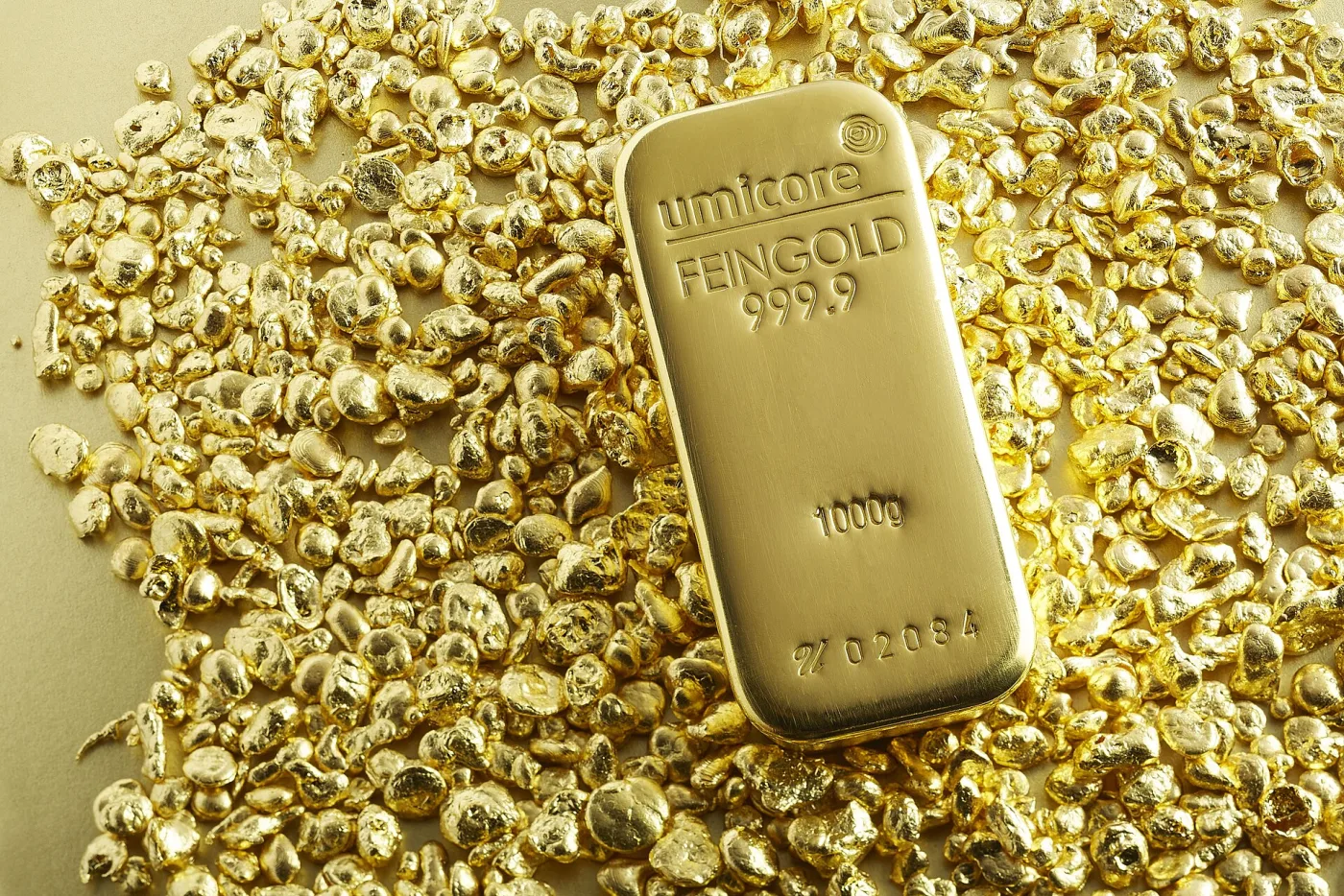 A gold bar with rounded corners lying on gold granules, stamped with the Umicore logo, FEINGOLD 999.9, 1000g and the serial number 02084