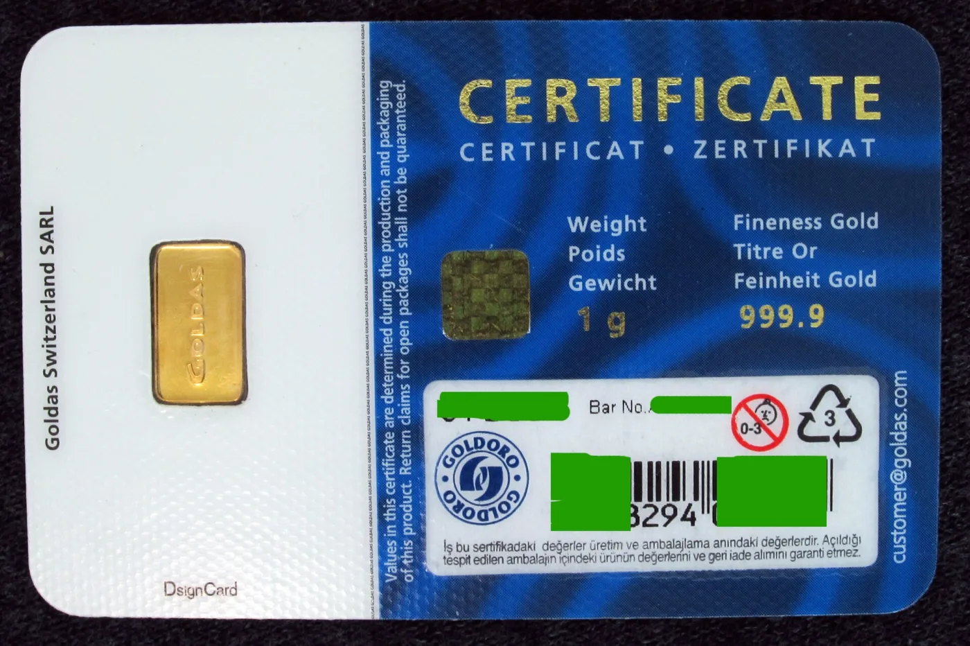 A blue and white certificate card with a tiny gold bar sealed in a window on the left; the card reads Weight 1 g and Fineness Gold 999.9, with the bar number and barcode blanked out in green