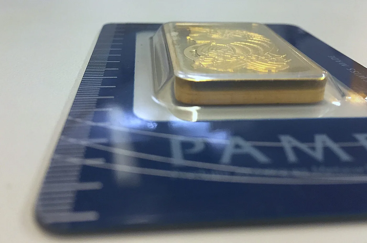 Side view of a gold bar sealed in a blister on a blue PAMP card, showing the bar's thick edge standing well above the card