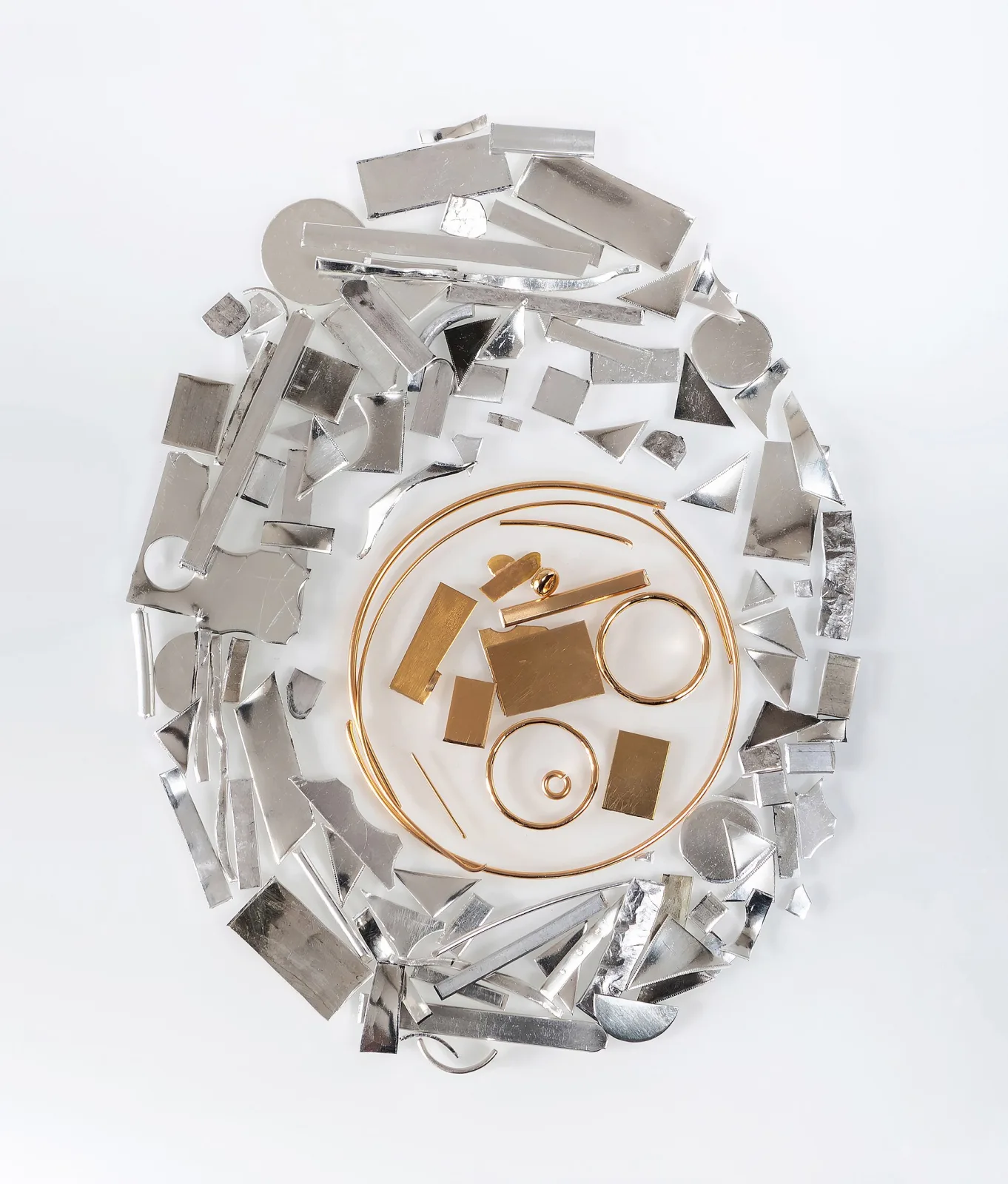 A pile of silver offcuts, wire and sheet on white paper with a smaller group of gold scrap pieces and two thin gold rings in the middle