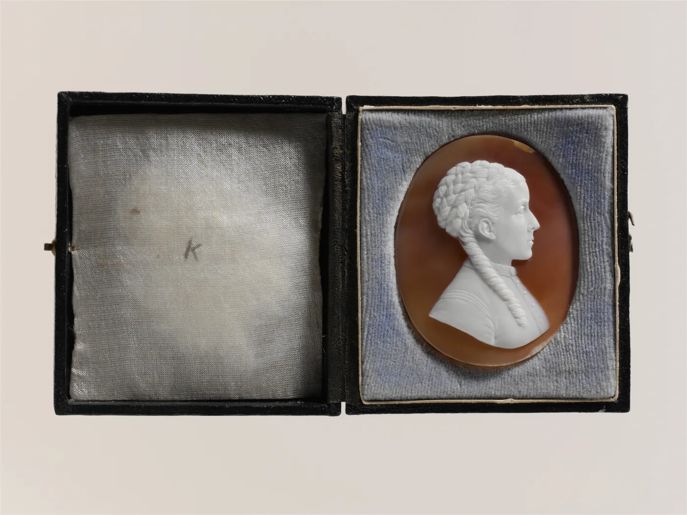 Shell cameo portrait of a young woman in right-facing profile, her hair braided into a crown with two long plaits down her back, wearing a high buttoned bodice, set in its original open leather case lined with blue velvet