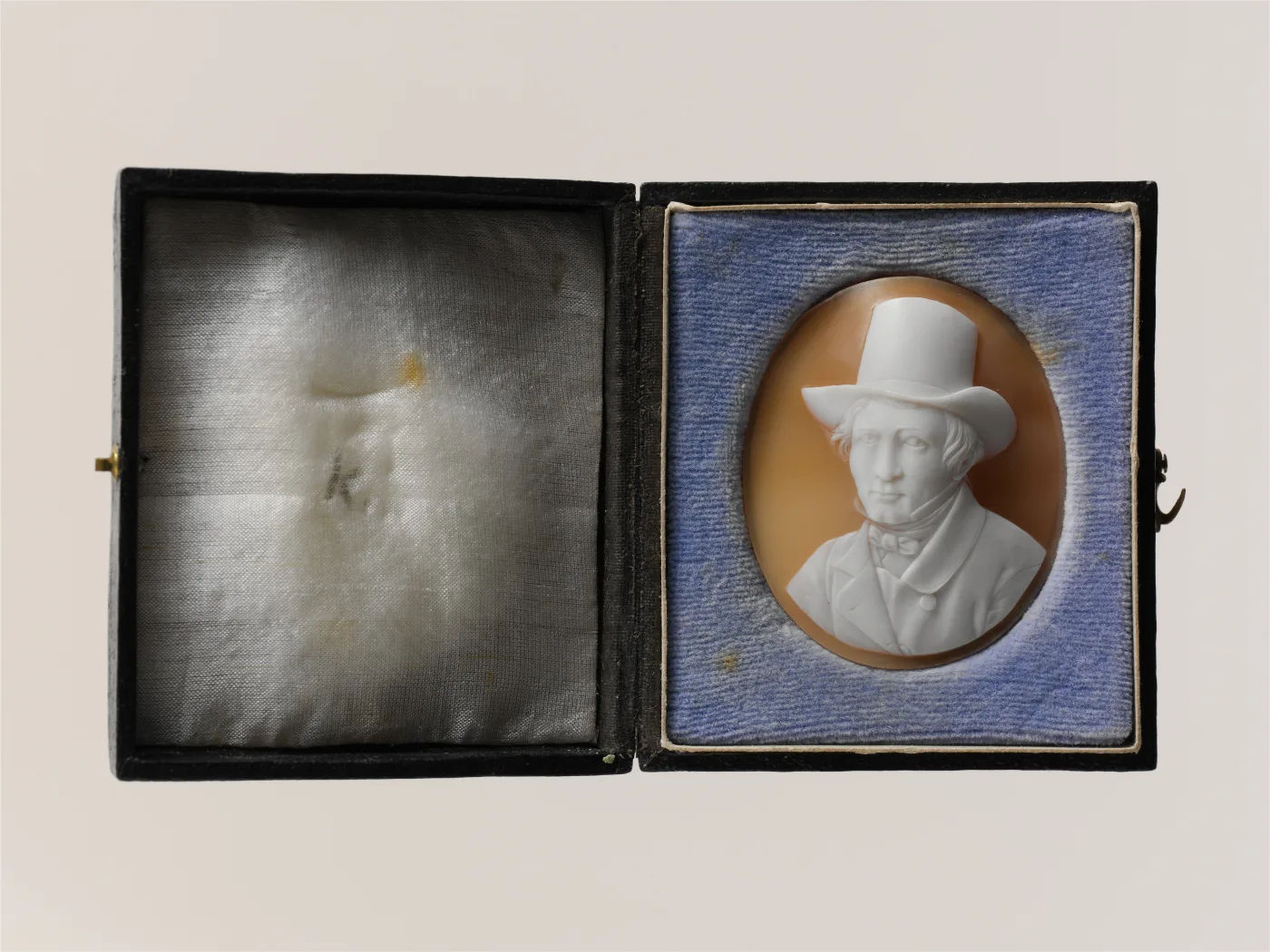 Shell cameo of an older man shown full face rather than in profile, wearing a tall top hat, a bow tie and a buttoned coat, in an open leather case lined with blue velvet