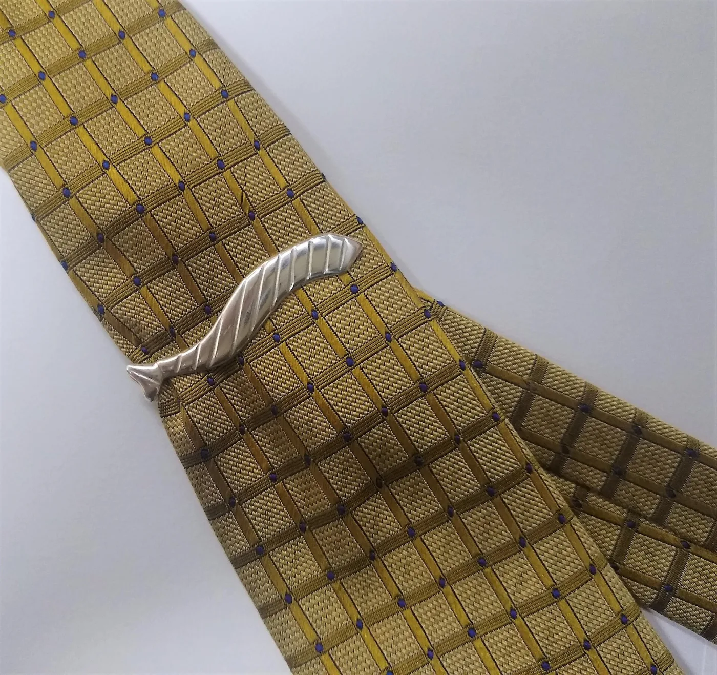 A silver tie clip with a sculpted wave-like end clipped to a patterned yellow and brown tie